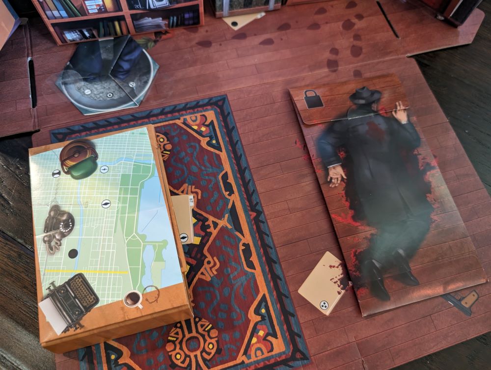 A birds-eye view of the floor of a 3D cardboard model of an office. On the left is a pop-up cardboard desk with a map and other desk items on top. On the right is a picture of a dead body lying face down and surrounded by blood. The picture of the dead body is printed on the outside of an envelope that matches the surrounding floor.