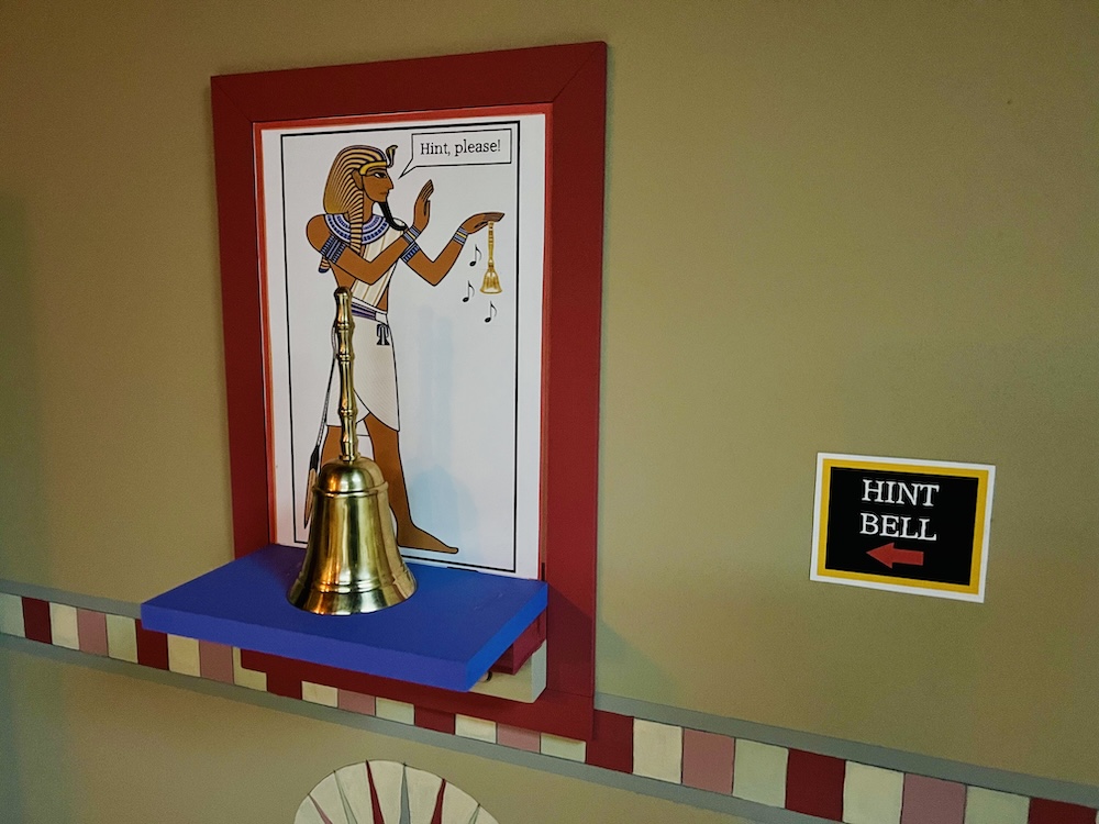 A golden bell on a shelf labeled, "Hint Bell." Behind it is a picture of a pharaoh holding a bell. A text bubble reads, "Hint please!"