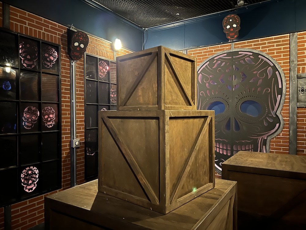 Day of the Dead themed game with skuls on the walls. In the middle of the room is a large stack of wood boxes.