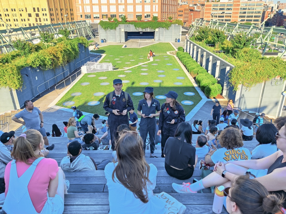 3 Great Gotham Challenge staff in blue coveralls and hats stand int he amphitheater of the Highline in NYC.