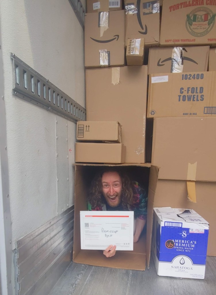 Matthew Stein on in a box in the back of a panel van filled with cardboard boxes.