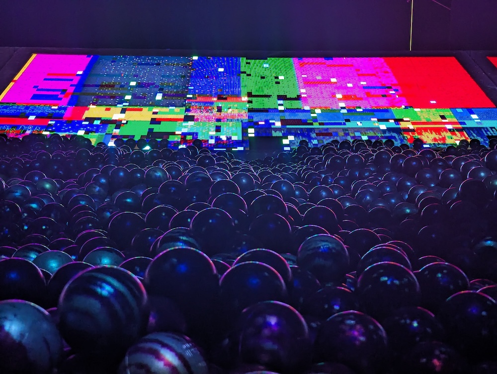 Rendering of a large ball pit with a strange blocky, multicolored, light display in the distance.
