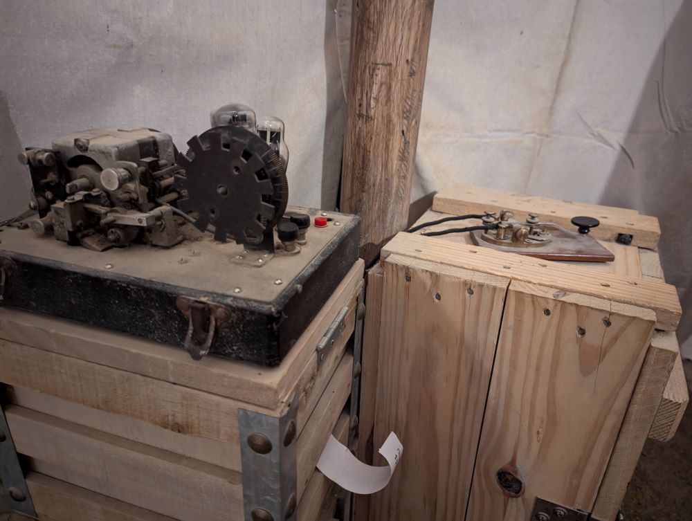Two wooden crates supporting an antiquated telegraph machine from the 1910s. The portion of the machine on the left crate has a large gear-like component, and a paper message curls out from the base of the crate. The right crate supports a telegraph tapper.