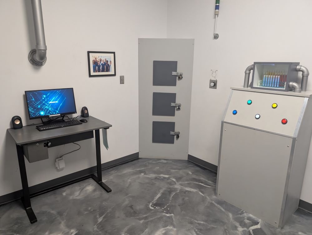The corner of a sparse and sterile lab. From left to right are a computer monitor on a small desk, a photo of some scientists, a corner cabinet with three locked doors, and a chest-height pedestal with five colored buttons and some colored test tubes on top.