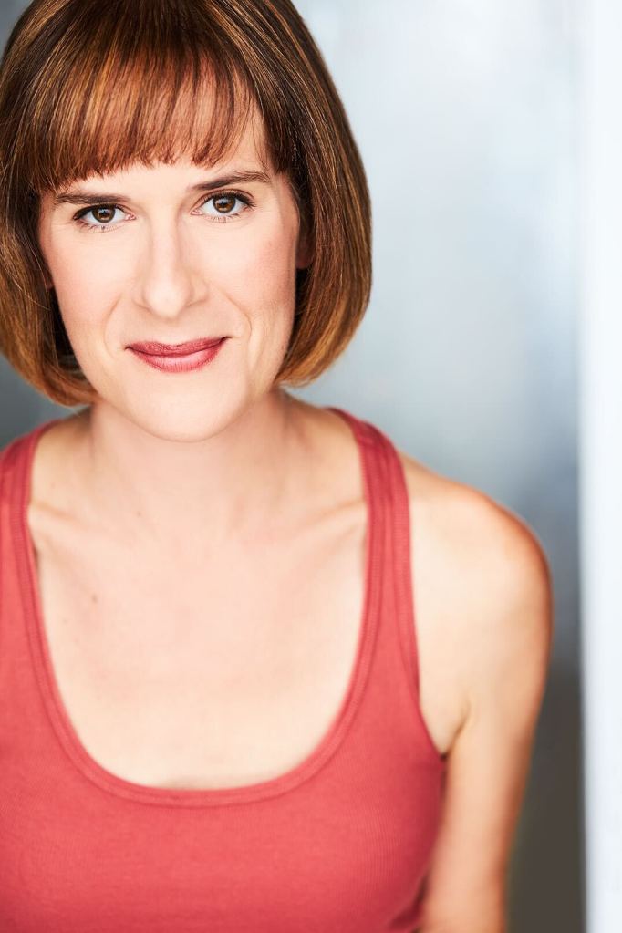 Woman with golden brown hair in a bob with bangs, smiling, wearing a pinkish red tank top