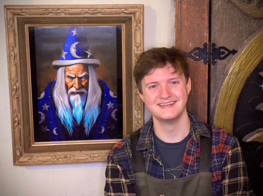 framed portrait of an old wizard in blue hat and robe covered with stars and moons. there is a man smiling next to it, clean shave, brown hair wearing a woodshop apron over a plaid shirt.