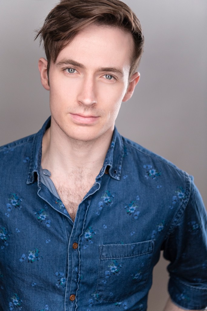 man with side parted hair, blue eyes, clean shaven, in a dark denim shirt with small flowers, partially unbuttoned at the collar.