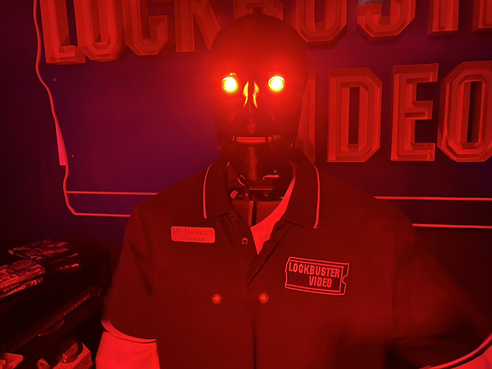 A robot Lockbuster Video employee standing behind the counter. Its eyes glow red. Its ID badge reads, "Mr. Manager, Manager"