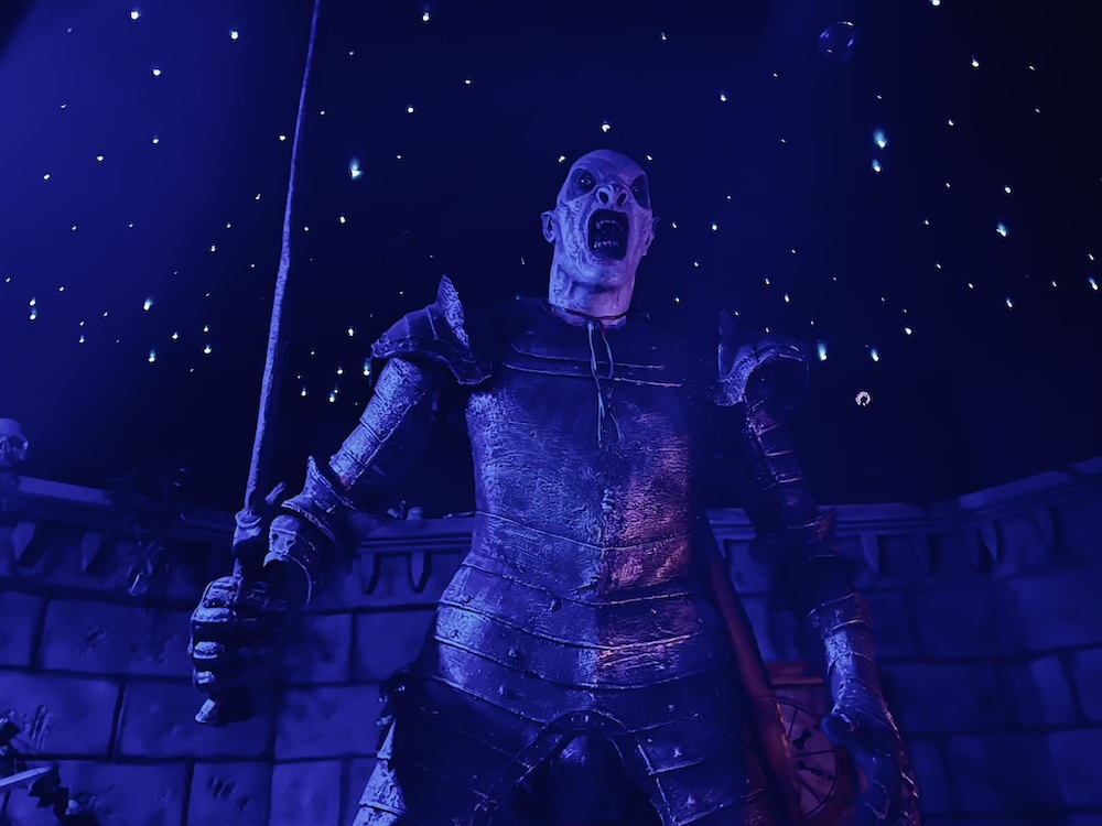 An ogre like character in a suit of of armor, holding a sword before a castle wall on a starry night.