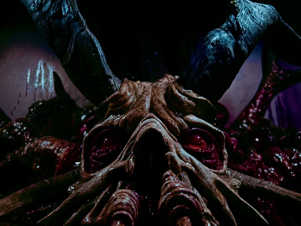 Closeup of the red-eyed, black horned, face of a demon. It looks wet and menacing.