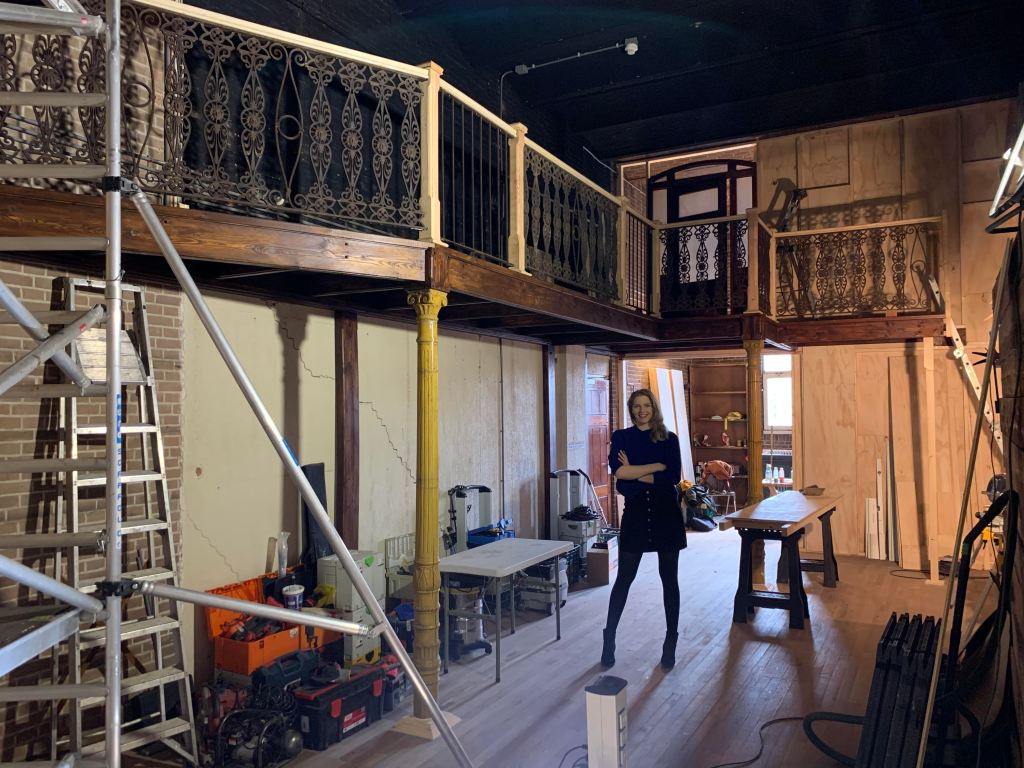 blond woman in blue sweater and black skirt standing in the middle of a room with a wrought iron balcony wrapping around. the room is partly built.