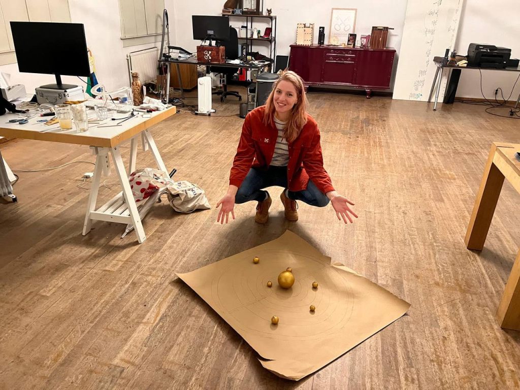 blond woman in orange jacket squatting in front of golden balls surrounding concentric circles, background of workshop office