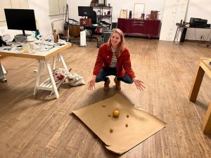 blond woman in orange jacket squatting in front of golden balls surrounding concentric circles, background of workshop office