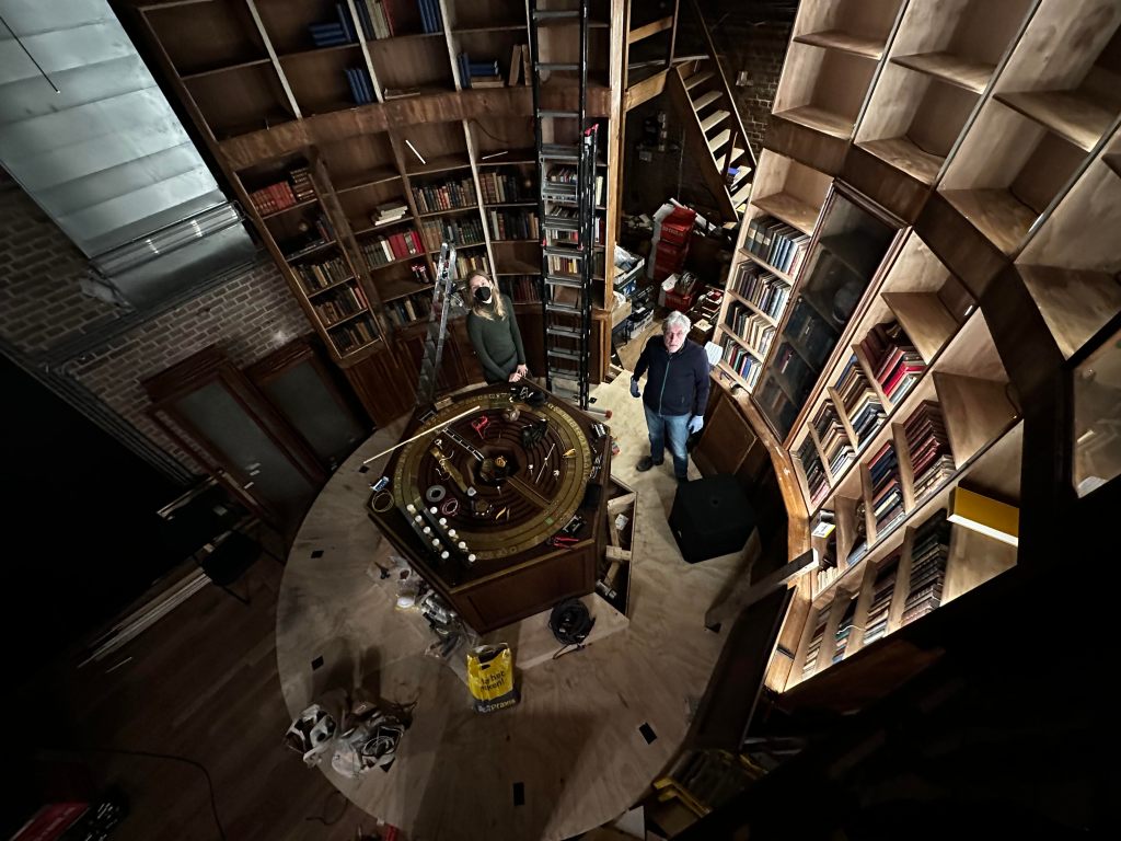 top down view of circular room with tall bookshelves and hexagonal table with two people standing looking up