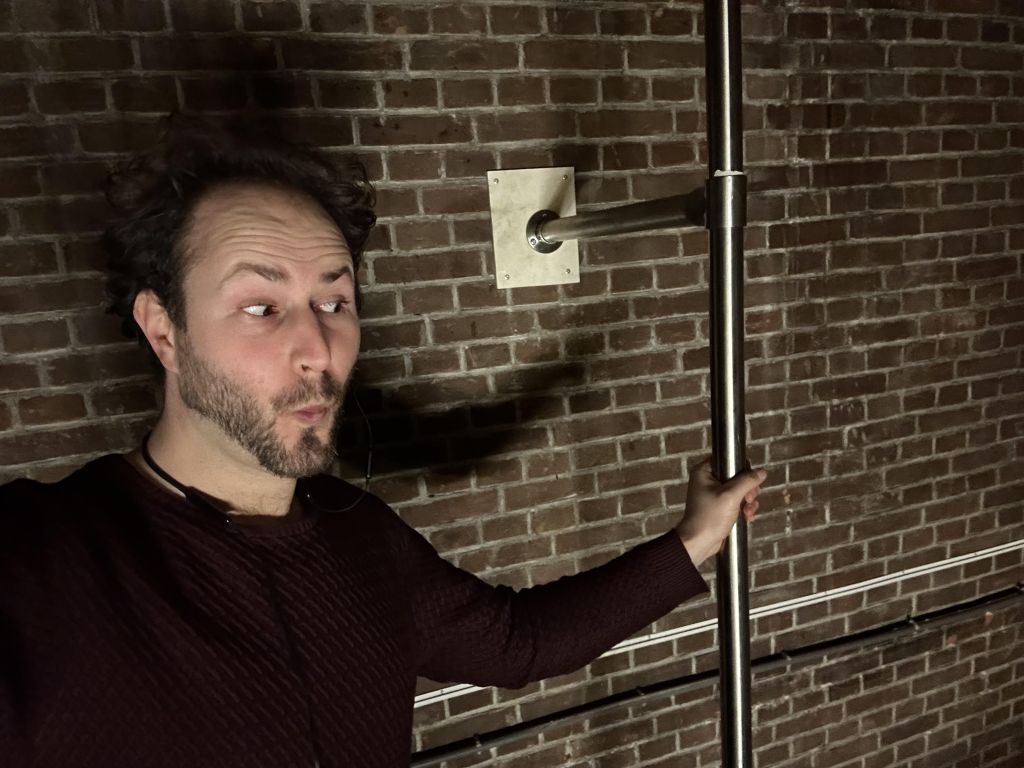 man with light brown hair and short beard in maroon sweater holding onto a vertical pole attached to a brick wall