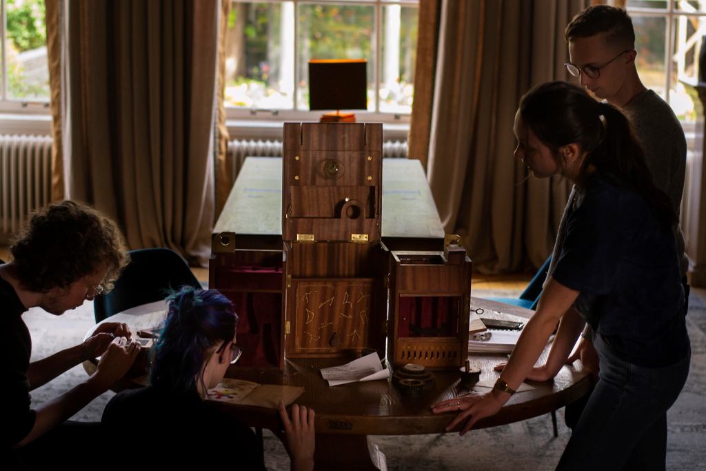 people standing around a wooden puzzle box that is open to reveal velvet compartments and golden constellations.