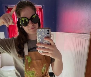 Audrey in a green slime-covered apron and goggles in a bathroom.