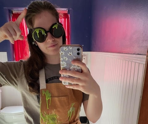 Audrey in a green slime-covered apron and goggles in a bathroom.
