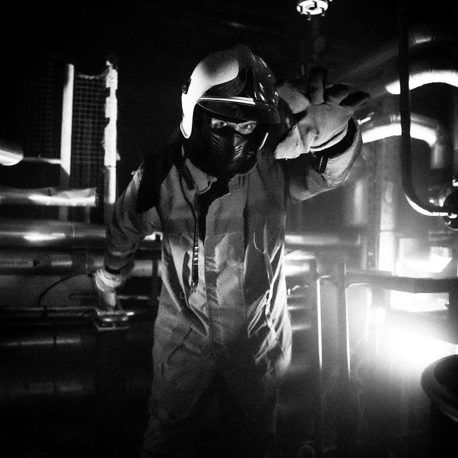 Marc dressed in coveralls, a helmet, mask and gloves reaching out towards the camera in an industrial environment.