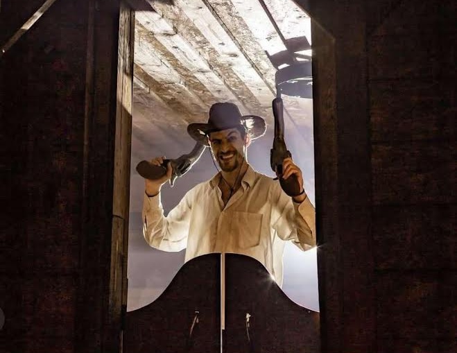 Philip dressed as an old west outlaw holding two cartoonish 6 shooters as they walk through the dramatically lit entrance to a saloon.
