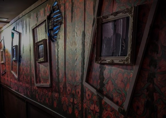 Three portraits hang along a dimly lit wall of red patterned wallpaper. Each portrait has an extra, oversized frame hanging at an odd angle around it.