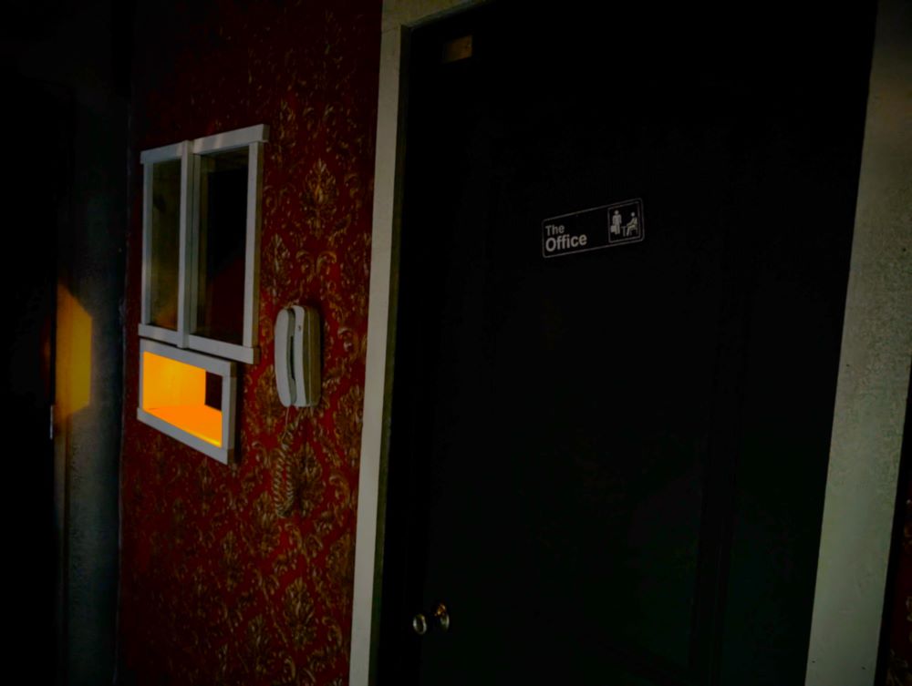 The dimly lit reception area of a motel lobby. To the left, a small sliding window appears above a brightly lit slot presumably for delivering keys to guests. A corded white phone hangs to the right of the window, and a closed door labelled as "The Office" appears to the right of the phone.