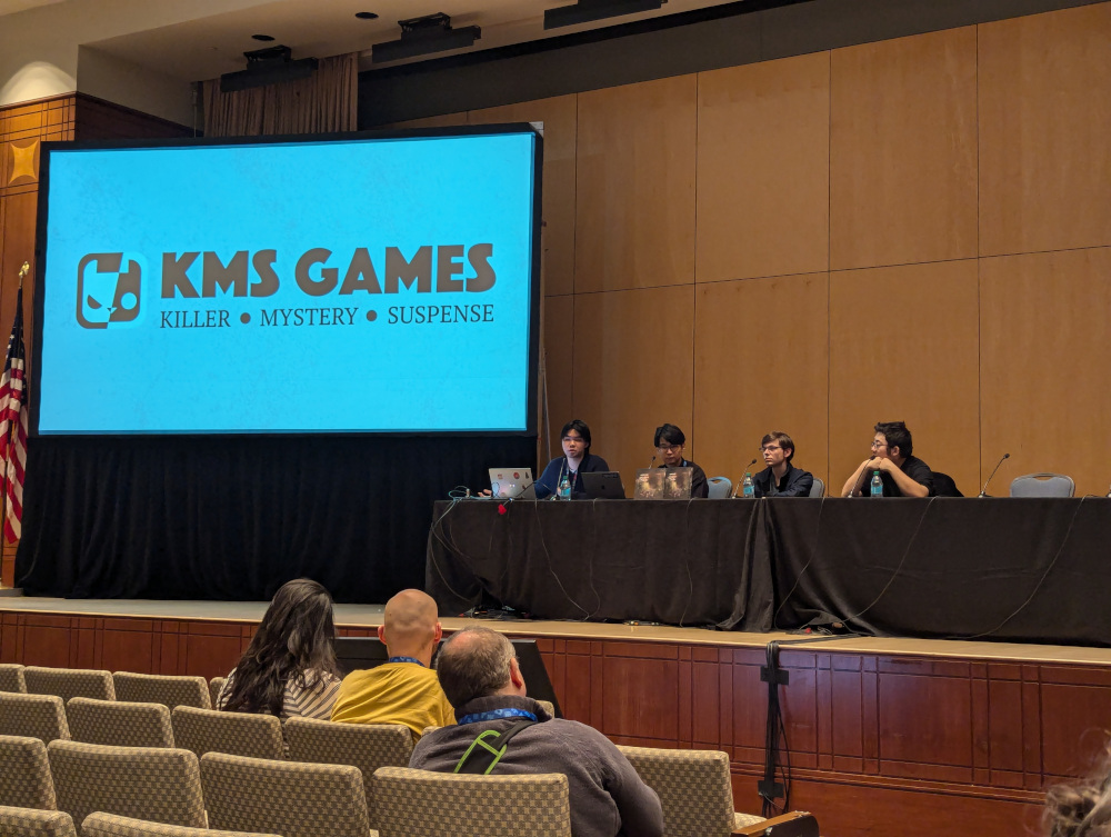 Four panelists sit at an elevated table. A large screen displays the logo for KMS Games.