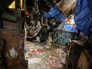 The interior of a cave made of garbage, the floor is covered in newspaper and product packaging, beyond, a car door, a lamp, and a tarp are in view.
