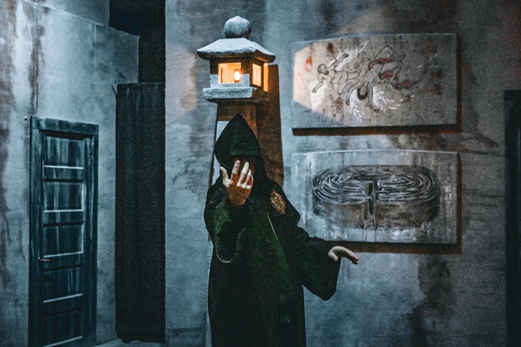 A black cloaked person reaching towards the camera, beckoning the viewer as they stand in a marble sanctum.