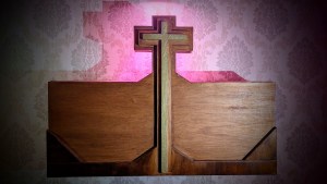 A wood cross in front of some old, floral wallpaper.