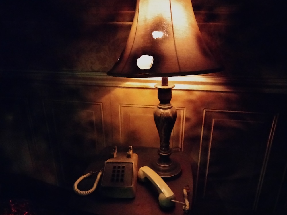 In a darkened room, a lamp with a singed shade sits on a small table next to a phone that's off the hook