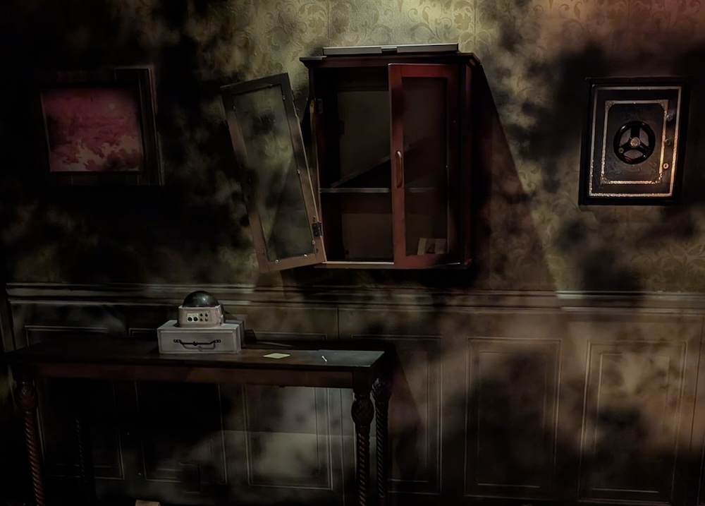 A framed picture, a cabinet with one door hanging open, and a safe on the wall above a table in a darkened room
