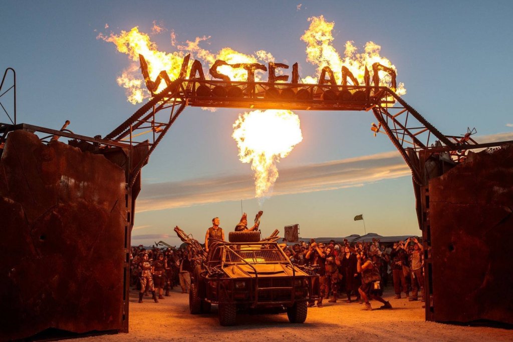 gate with large sign that reads "wasteland". the sign is on fire. There is a modified jeep below surrounded by people in dystopian costumes.