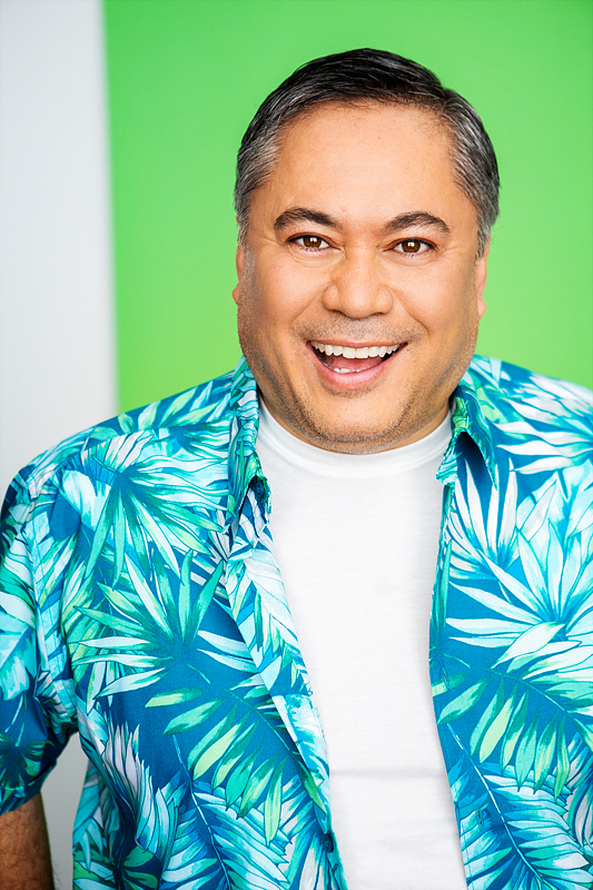 filipino man, clean shaven, smiling wearing a white t-shirt and blue hawaiian shirt unbuttoned over that.