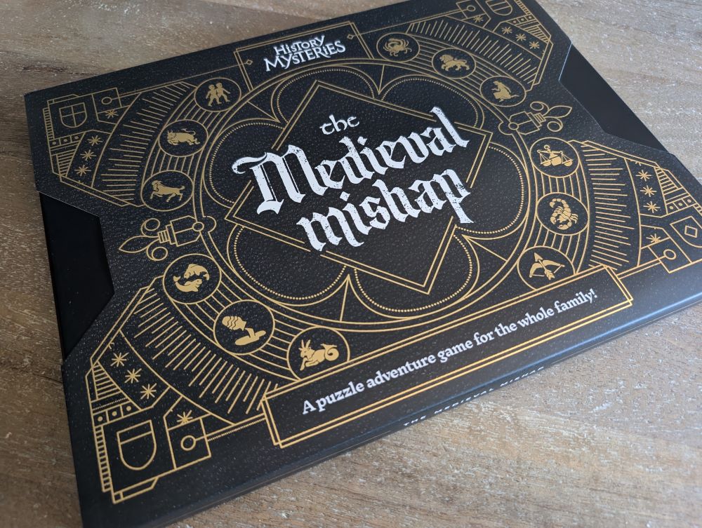 A photo of the rectangular game box for The Medieval Mishap. The box is black with an ornate golden design and white lettering. The gold design is mostly symmetrical line art that includes iconography of shields and zodiac symbols.