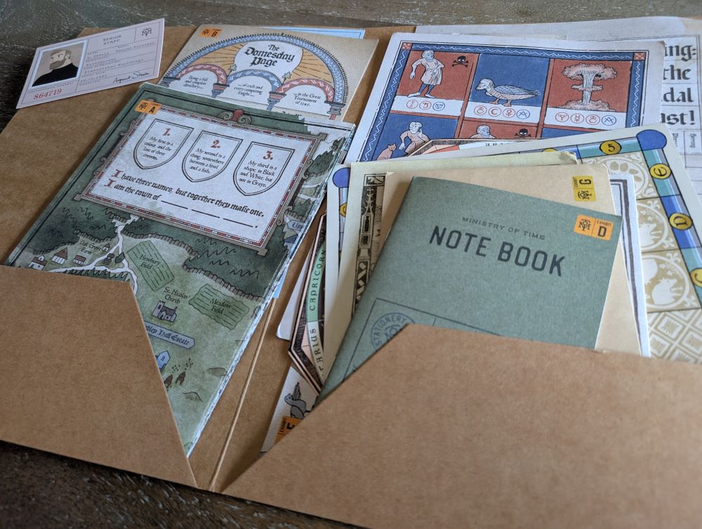 A photo of an open brown folder stuffed full of various colorful game materials. In the left pocket there are an identification card for a time traveling agent, a folded map, and something called "The Domesday Page." In the right pocket is a small green note book stacked atop several unidentifiable but colorful documents.