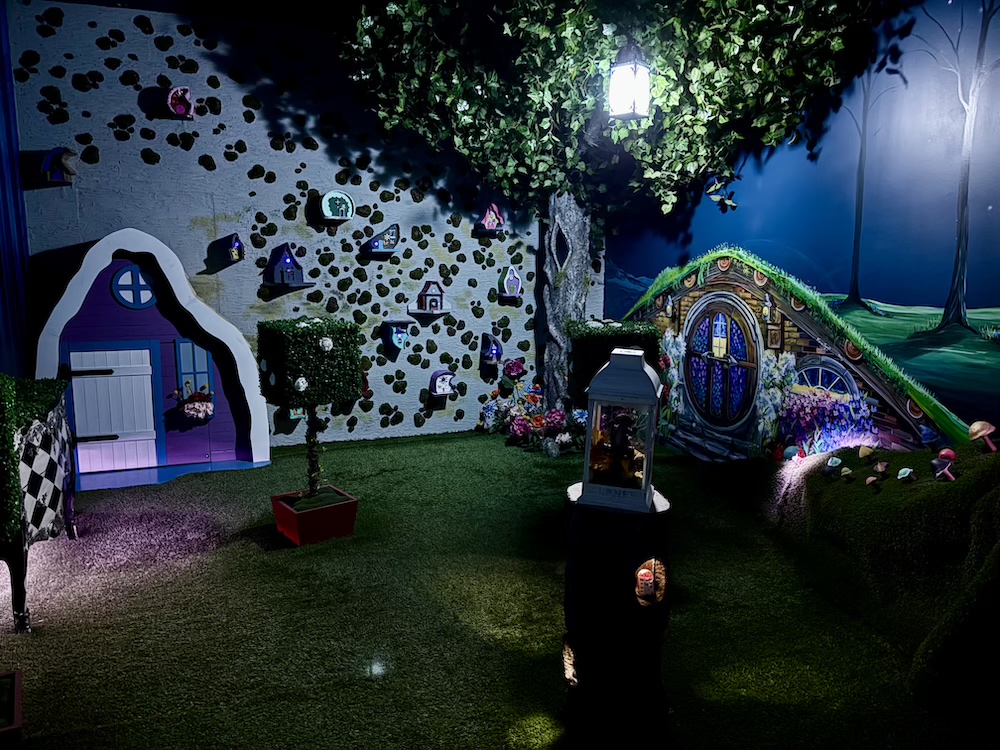 A wonderland set with a small house, a wall of clocks, and a tree with a lantern hanging from a branch.