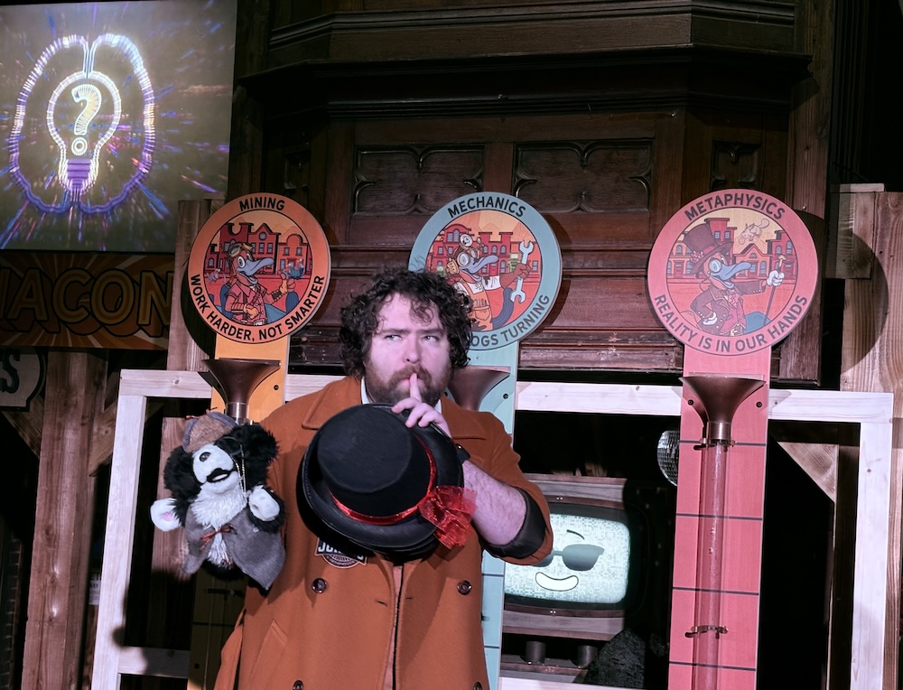 A beared character from Phantom Peak wearing a trench coat with a detective dog puppet on his hand, holding his other finger up to his mouth in a "shhh" gesture.