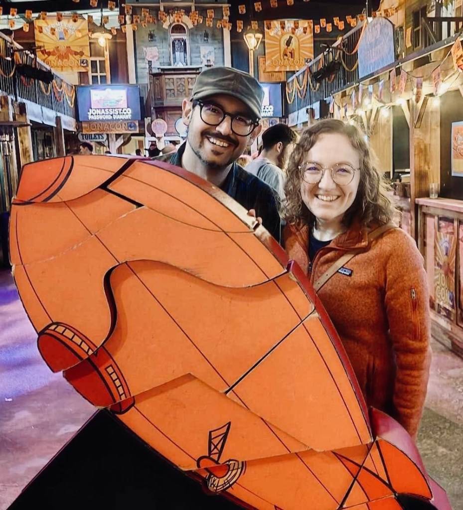David and Lisa standing behind a large, vertical, orange blimp jigsaw puzzle in the middle of the Phantom Peak town square, surrounded by stalls.