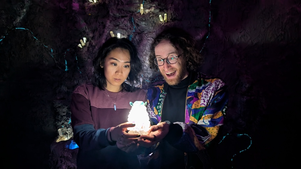 A man and a woman in an enchanted cave. Together they are holding a glowing gemstone and staring at it with awe.