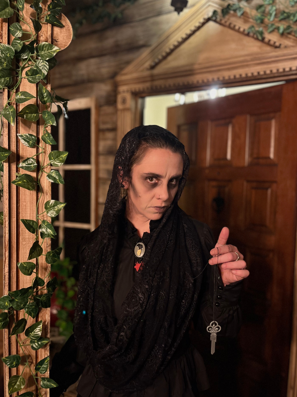 Woman with ghostly makeup wearing a black dress and head veil, holding an ornate key on a string and standing in front of a slightly open door.