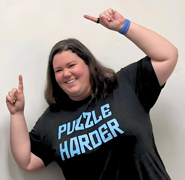 Woman with shoulder length brown hair parted on the side wearinga black t-shirt that says "puzzle harder". both arms in the air, she is leaning to the left and pointing upwards.