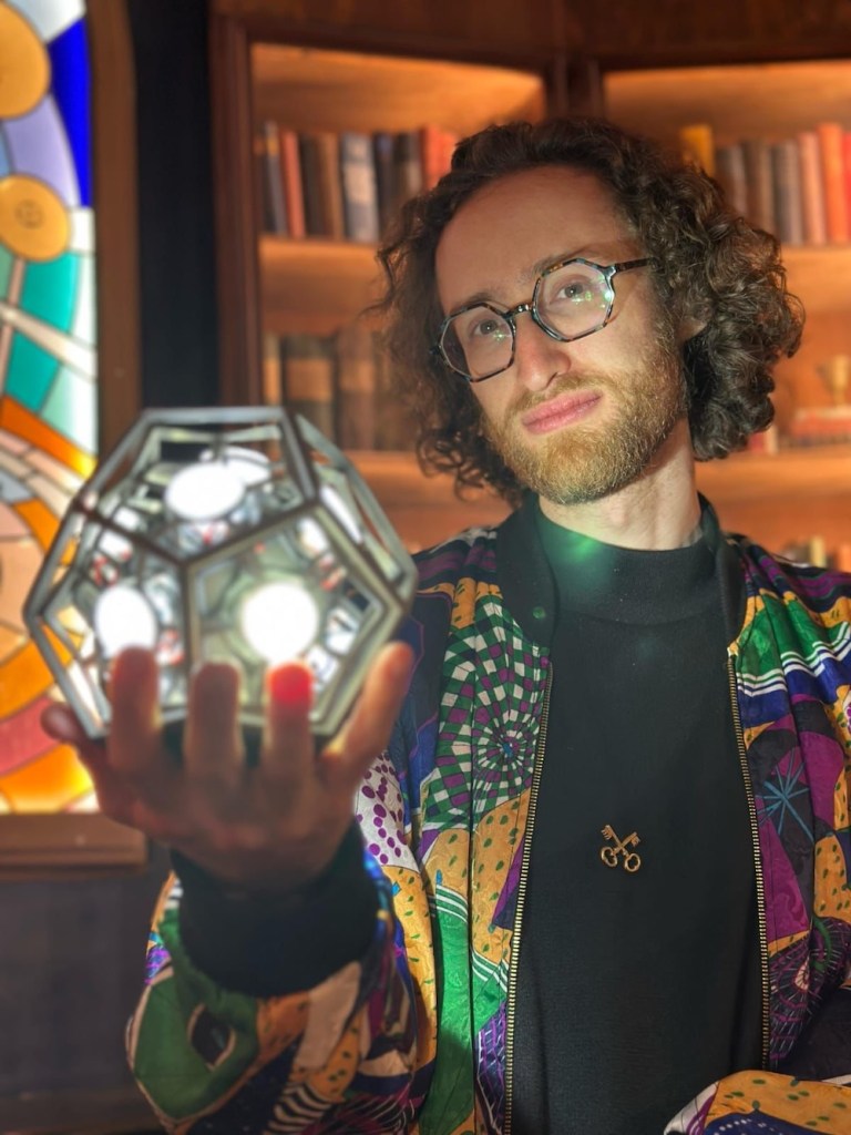 A beared man with glasses standing in a library-like space holding a glowing geometric sculpture out in his hands.
