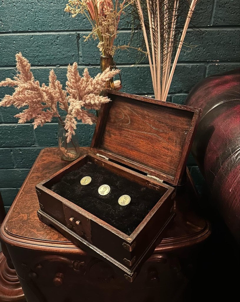 An elegant wooden box holding 3 metal pins, on a side table beside dried flowers.
