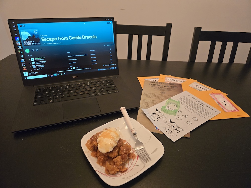 Escape From Castle Dracula's spotify playlist open on a computer, beside the games's paper puzzle components, a set of plastic fangs, and a plate with an ice cream dessert.