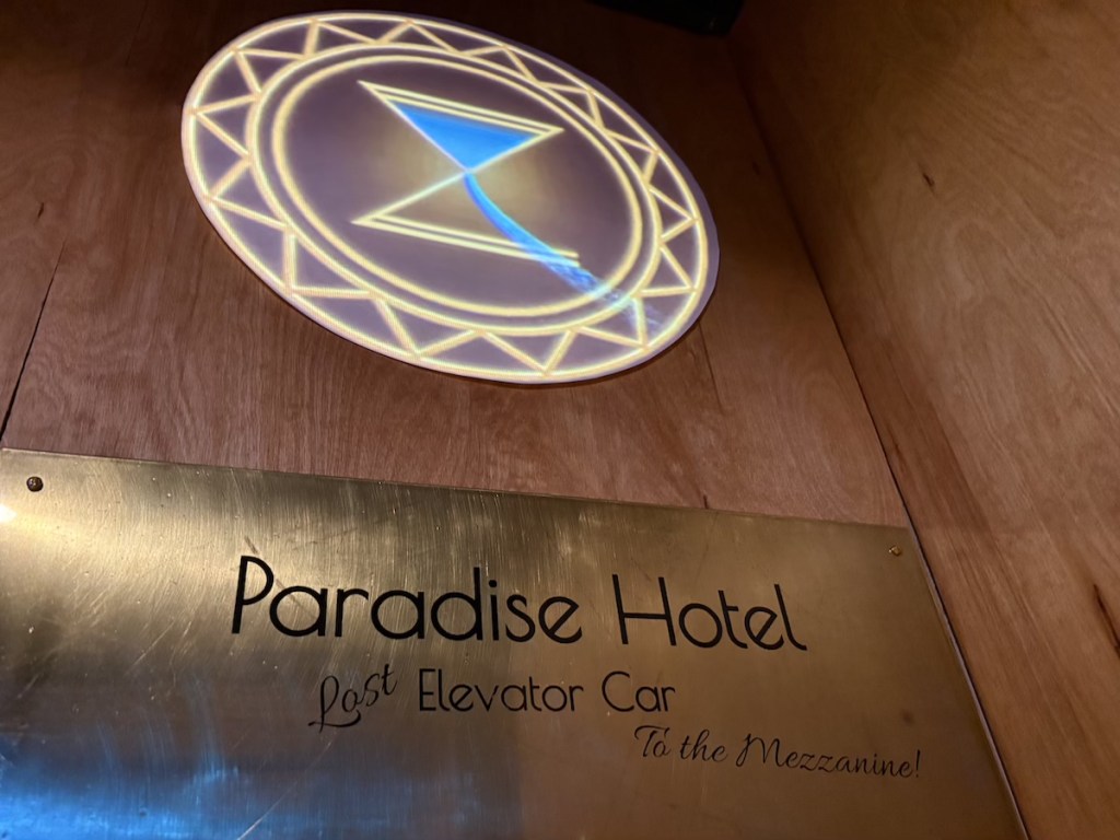An ornate leaking hourglass logo above a plate that reads, "Paradise Hotel Last Elevator Car to the Mezzanine."