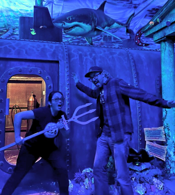 asian woman wearing glasses, black logo t-shirt and jeans wielding trident at man in black cap, plaid shirt and jeans. they are standing in an underwater set with a shark and submarine in background