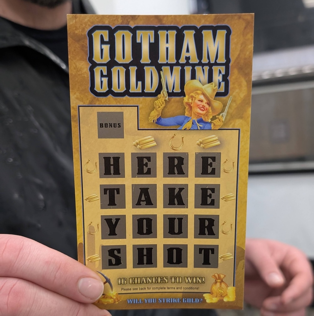 A scratch off ticket with a cowgirl holding two guns, it's titled, "Gotham GoldMine." The grid of scratch-offs reads, "HERE TAKE YORU SHOT"