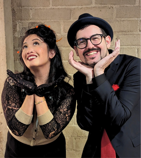 asian woman in lace and beige cardigan with net veil. man with fedora, glasses, in black suit with red tie. Both have hands cupped under their chin