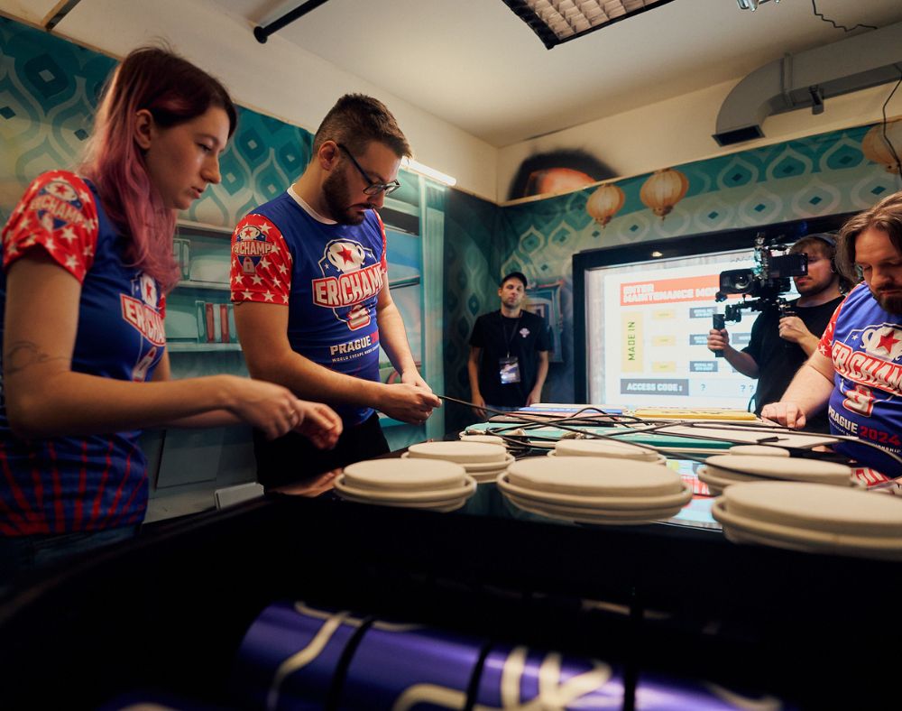 Three players in matching jerseys working on an escape room puzzle while being filmed and observed during the 2024 Escape Room Championship